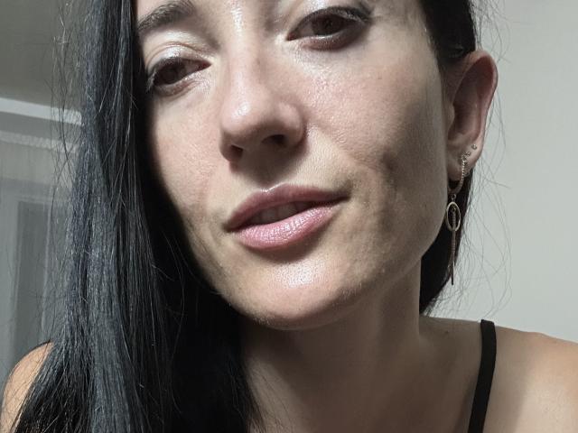 BellaJulls with shiny skin, black straight hair, small hoop earrings, and subtle smile, wearing a black strap top, showing her nose, lips, and slightly upturned eyes.