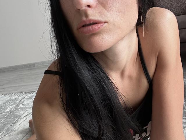 BellaJulls has black hair, light skin, and is wearing a black tank top. Her pouty lips are slightly parted, and she's looking down with a soft expression.