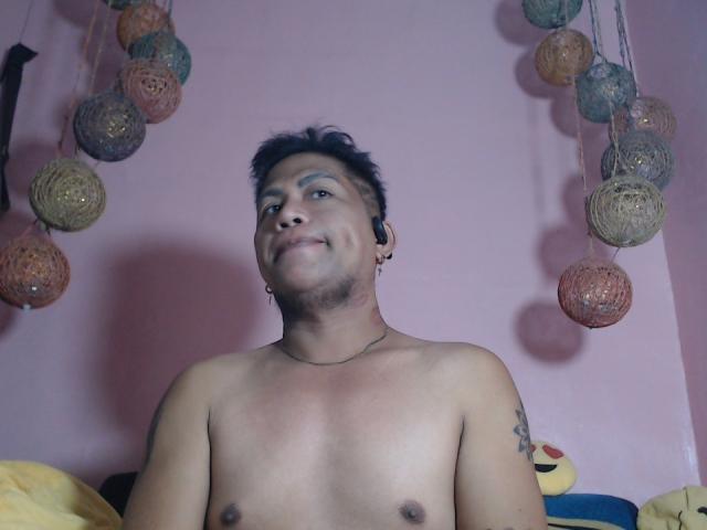 Shirtless BigcockPinoyCumback with short black hair, small tattoo on right shoulder, ear piercings, smiling, hanging colorful balls in pink room, visible yellow pillow, slight tan skin.