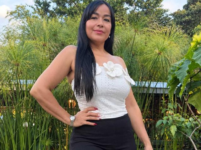 TamaraFoxy, with long black hair, wearing a white off-shoulder top with flower details, black skirt, and wristwatch, smiling, hand on hip, standing in front of greenery.