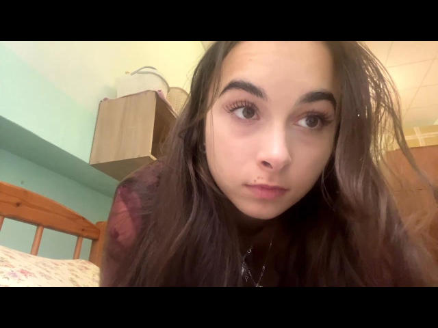 DonyaDuraku with long brown hair, big brown eyes, and a neutral expression, wearing a dark shirt, looking at the camera, in a slightly messy room with a wooden bed.