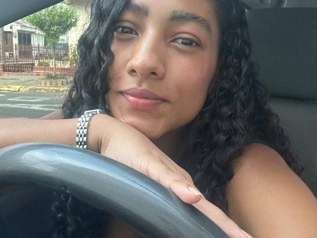 NatalyBlonder with curly black hair, medium brown skin, and green eyes, holding the car's steering wheel, wearing a silver watch, and giving a slight pouty smile.