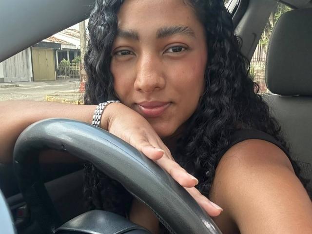 NatalyBlonder with curly black hair, tan skin, and brown eyes, leaning on the car steering wheel, wearing a black top, and a silver bracelet, giving a subtle smile.