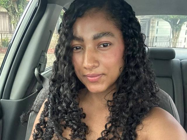NatalyBlonder with curly black hair, medium brown skin, and pink lips, sitting in a car, wearing a grey top, looking at the camera with a slight smile.