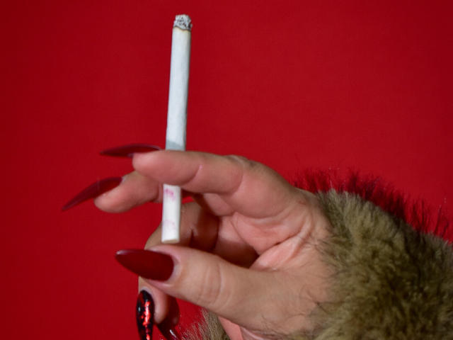 La main de VanesaMoka, avec de longs ongles rouges et du vernis noir, tient une cigarette sur un fond rouge, et elle porte un bracelet marron en fourrure. Les seins ne sont pas visibles..