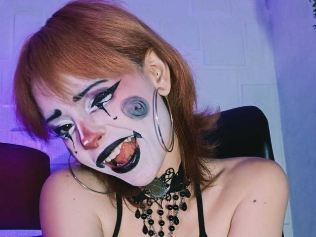 ViolettClay with painted white face, black eyeliner, gray eye makeup, red nose, black lipstick, hoop earrings, black choker, and red hair, showing a crazy grin, tits visible.