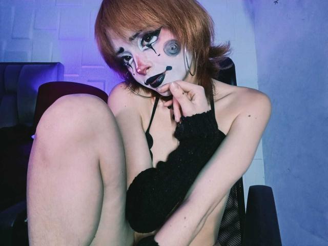 ViolettClay, à la peau pâle et aux cheveux roux, porte du maquillage sur le visage, du rouge à lèvres noir et un soutien-gorge noir, est assise sur une chaise, les genoux repliés, a un manchon noir, et pose un doigt sur son menton, avec un éclairage bleu..