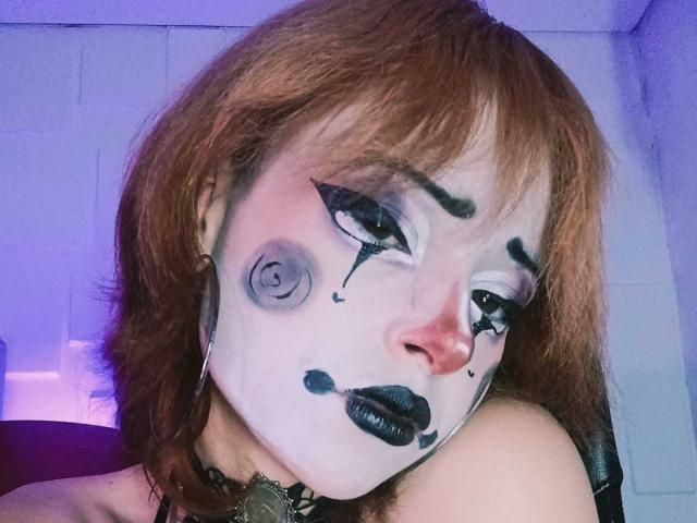 ViolettClay with painted white face, black eyeliner, and dark lipstick, has red hair, black eyeshadow, and gray eye makeup, wearing a black choker, in a dimly lit room.