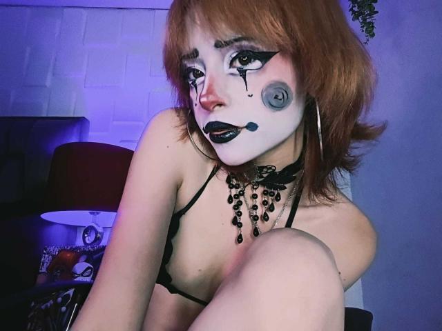 ViolettClay, con maquillaje blanco en la cara, lápiz labial negro y una nariz roja, lleva un sujetador negro y un collar de cuentas, con las rodillas levantadas, mostrando sus pequeños pechos. De cabello pelirrojo, luce seductora..