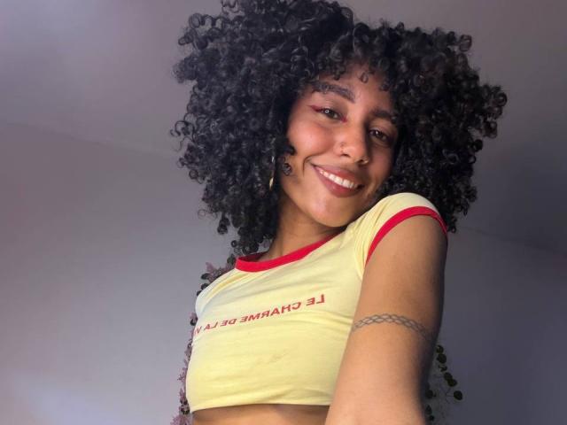CinthiaWalker with curly black hair, smiling, wearing a yellow crop top with red trim, showing her small tits, tattoo on right arm, and dark skin.