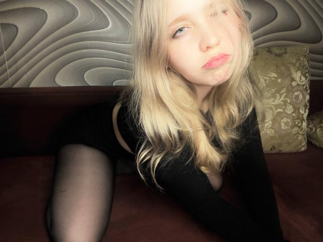 TiffaniMacandog with blonde hair, pouty pink lips, and black outfit, leaning on a red couch, wearing sheer black stockings, her tits slightly visible through the top.