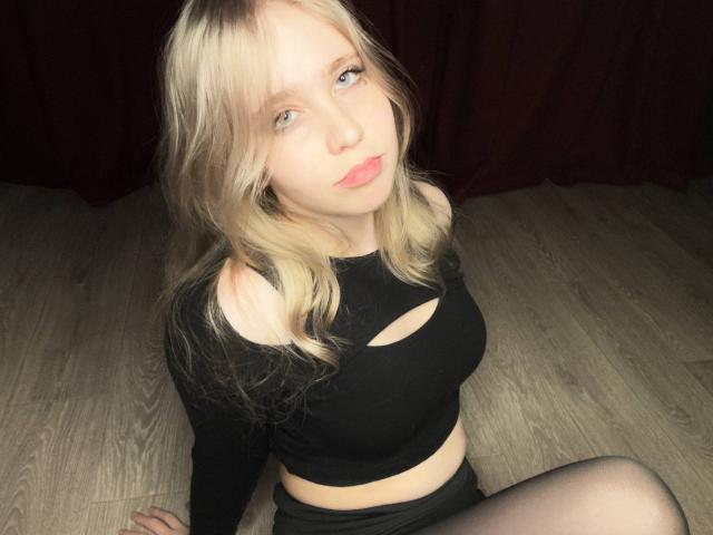 TiffaniMacandog with blonde hair, black cutout top, and black tights, sitting on wooden floor, looking up with pink lips, pale skin, and blue eyes. Boobs visible through top.
