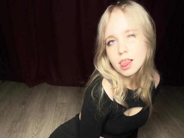 TiffaniMacandog with blonde hair, winking, sticking out her tongue, wearing a black top with a cutout, on wooden floor, dark red curtain in background.