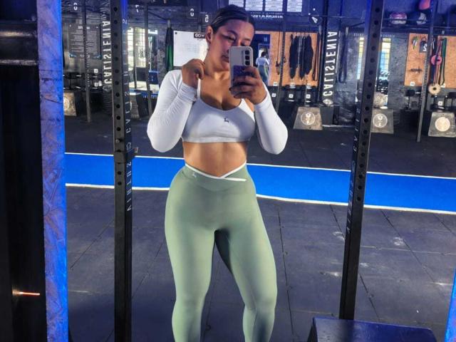 EmaFoxy, con sus pechos a la vista en un top corto blanco, sosteniendo su barbilla, usando leggings verdes de cintura alta, tomando una selfie en un espejo en un gimnasio..