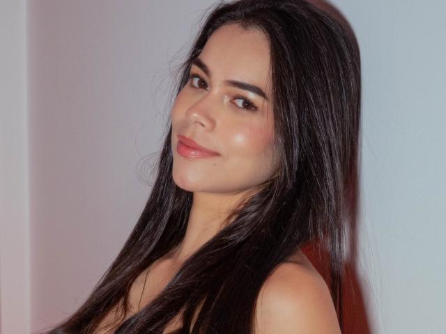 Alana Blake, con su cabello negro largo y lacio, ojos marrones y una sonrisa sutil, muestra sus hombros desnudos y un ligero atisbo de sus senos, todo ello bajo una luz suave y natural..