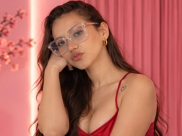 SkinnySweetMilff with glasses, red dress, small heart tattoo on shoulder, hand on cheek, gold earrings, pink background, long brown hair, pouty lips, looking serious, sexy, cute.