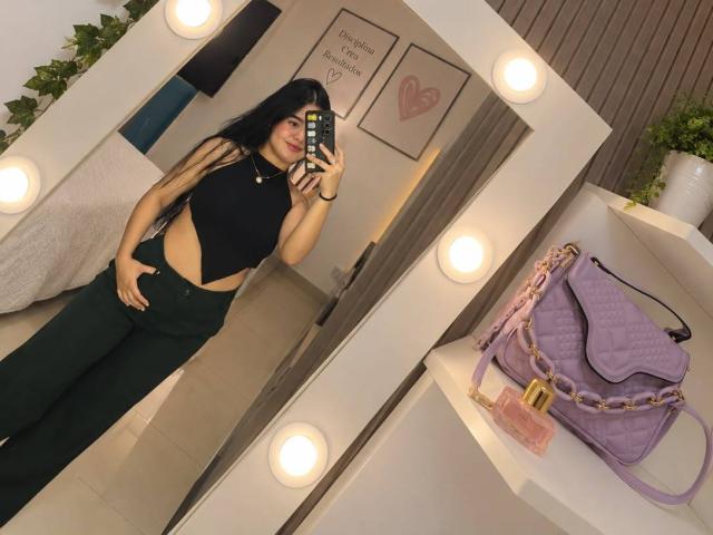 MalloryRamos has long black hair, wearing a black crop top and green pants, showing a cutout midriff, standing in front of a mirror with round lights, holding a phone.