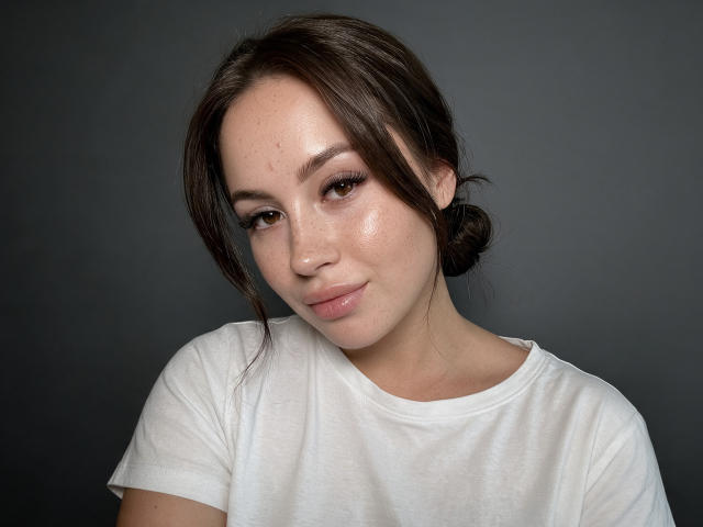 NensySoterr has shiny skin, brown eyes, and a subtle smile. She wears a white shirt, with dark brown hair in a loose bun. Her small tits are slightly visible.