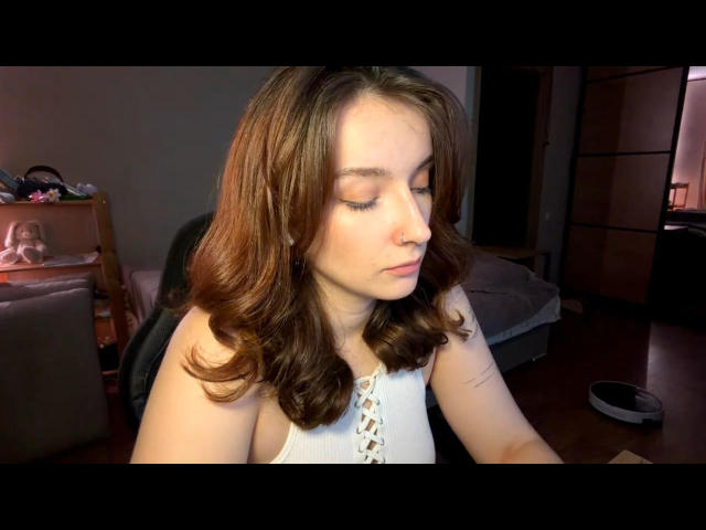 ConsueloKissam with brown wavy hair, nose ring, and white lace-up top, looking down, has small tits and a tattoo on her right arm, sitting in a dimly lit room.