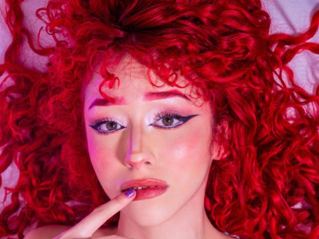 GaiaDoll with red curly hair, pink lipstick, and blue eyeshadow, lying down, touching her pink lips with a finger, looking at the camera, with a sexy, seductive expression.