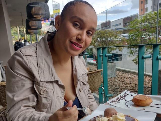 TanyaMartin with short hair, pink lipstick, and tan jacket, holding fork, smiling, eating dessert, donut on plate, small chocolate, outside on green metal balcony, overcast sky.