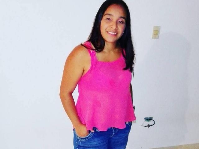 TanyaMartin with medium brown skin, long black hair, pink semi-sheer tank top, blue jeans, smiling, hands in pockets, standing against white wall, light switch to right.