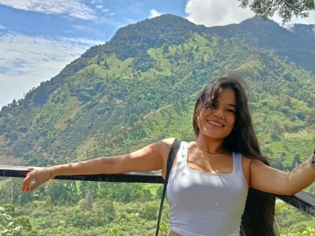 Isabella Voss, con lunghi capelli scuri, sorridente, indossa una canotta bianca, con le braccia appoggiate a una ringhiera nera, mostrando il seno, in piedi di fronte a una montagna verde..