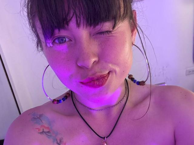 Iniridax, che fa l'occhiolino con il viso illuminato di viola, rossetto rosso, orecchini a cerchio e una collana nera, mostrando lentiggini e un tatuaggio colorato sulla spalla sinistra. Senza maglietta, con le clavicole visibili..