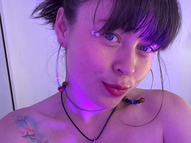 Iniridax, avec un visage illuminé de violet, des cheveux bruns, une frange, des boucles d'oreilles, un collier de perles colorées, du rouge à lèvres rouge, un tatouage sur l'épaule gauche, un léger sourire, les épaules dénudées, des taches de rousseur..