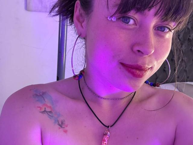 Iniridax has purple-tinged skin, dark brown hair, blue eyes, red lipstick, and a colorful tattoo on her shoulder. She's topless, wearing a necklace with a pink stone.