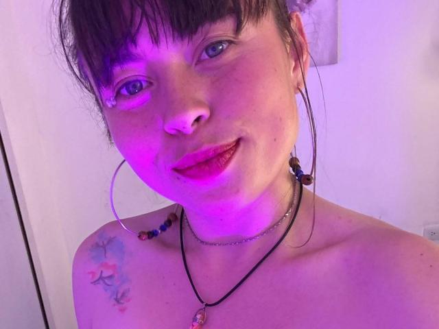 Iniridax, con il viso illuminato di viola, rossetto rosa, orecchini a cerchio, collana, tatuaggio sulla spalla, frangia e un leggero sorriso, che mostra le spalle scoperte. Iniridax ha gli occhi azzurri e la pelle chiara..