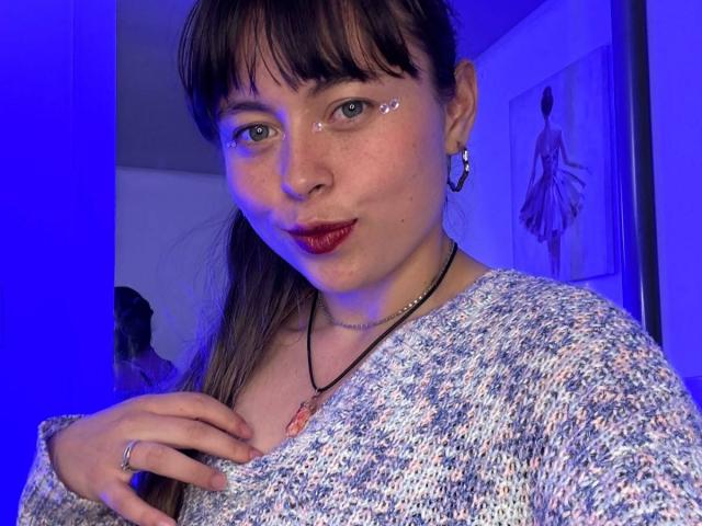 Iniridax with brown hair, bangs, and blue eyes, wearing a textured sweater, red lipstick, and a nose ring, has a small shiny sticker on her cheek, blue light.