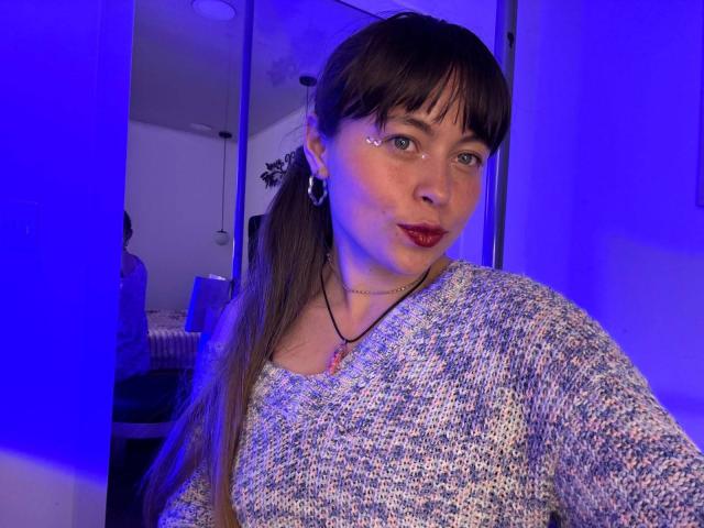 Iniridax with brown hair, bangs, and green eyes, wearing a blue and white textured sweater, has shiny face, red lipstick, and small hoop earrings, under blue lighting.