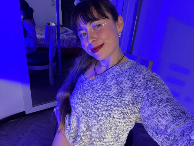 Iniridax mit langen, braunen Haaren, Pony und rotem Lippenstift, trägt ein graues, strukturiertes Oberteil, lächelt in die Kamera, im Hintergrund ist blaues Licht zu sehen, sie hält eine Halskette..