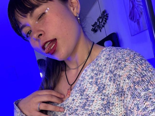 Iniridax with a snotty nose, tongue out, wearing a floral sweater, necklace, and earrings, blue lighting, hand on chest, ring on finger, and makeup on cheek.