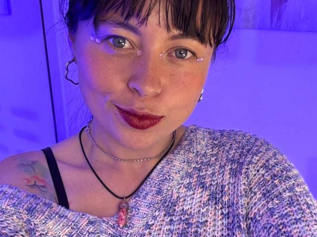 Iniridax with tear tattoos under eyes, wearing red lipstick, black strap top, gray textured sweater, black necklace with pink gem, and small tattoo on left shoulder, in purple light.