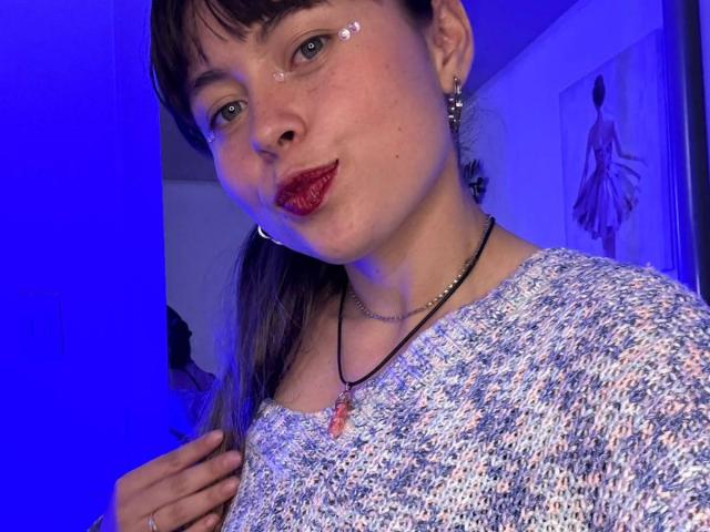 Iniridax has red lipstick, sparkly eye makeup, a speckled shirt, and a pink gem necklace, with blue lighting. Iniridax's hand touches her chest. Iniridax has dark hair in a ponytail.