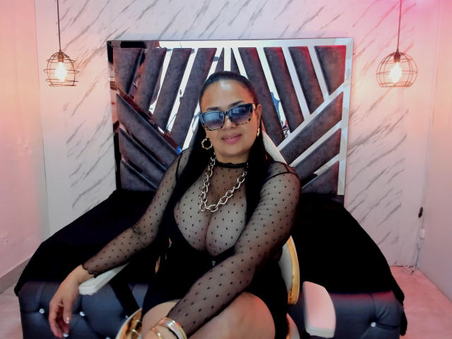 MiaaVelvett with big tits, wearing a sheer black polka dot dress, chain necklace, blue glasses, and hoop earrings, sitting on a black tufted chair, smiling.