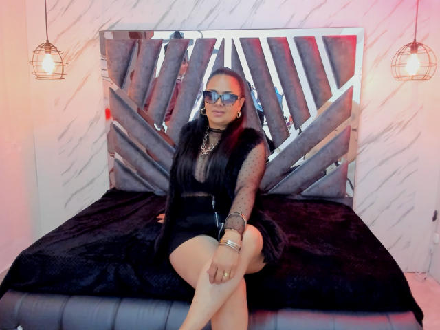 MiaaVelvett sits on a black bed, wearing a sheer black top, black shorts, and blue sunglasses, with a fur vest and jewelry, looking sexy with her tits visible.