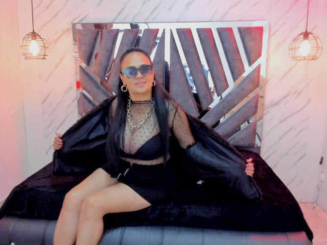 MiaaVelvett with long black hair, sunglasses, gold earrings, sheer polka-dot top, black bra, black fur jacket, black skirt, sitting on bed, holding jacket open, showing tits.