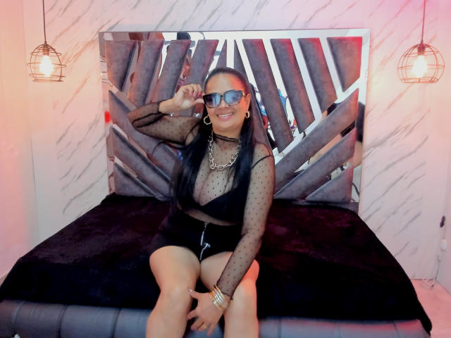 MiaaVelvett sitting on a bed, wearing a sheer black polka dot top, black shorts, and blue sunglasses, with long black hair, smiling, and holding her glasses.