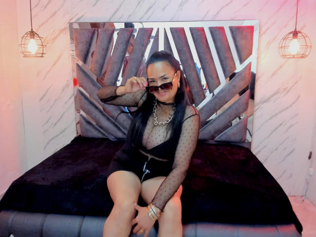 MiaaVelvett sitting on a bed, wearing a sheer black polka dot top, black shorts, large sunglasses, and a gold necklace, with her hand near her eye. Bedroom setting.