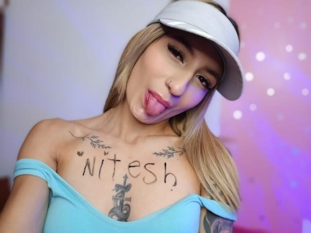 Kaiix, com cabelo loiro, viseira branca, blusa azul com um ombro à mostra, língua de fora, tatuagens no peito e no braço, língua rosa protuberante, pequeno piercing no nariz, tatuagens no osso do colo..