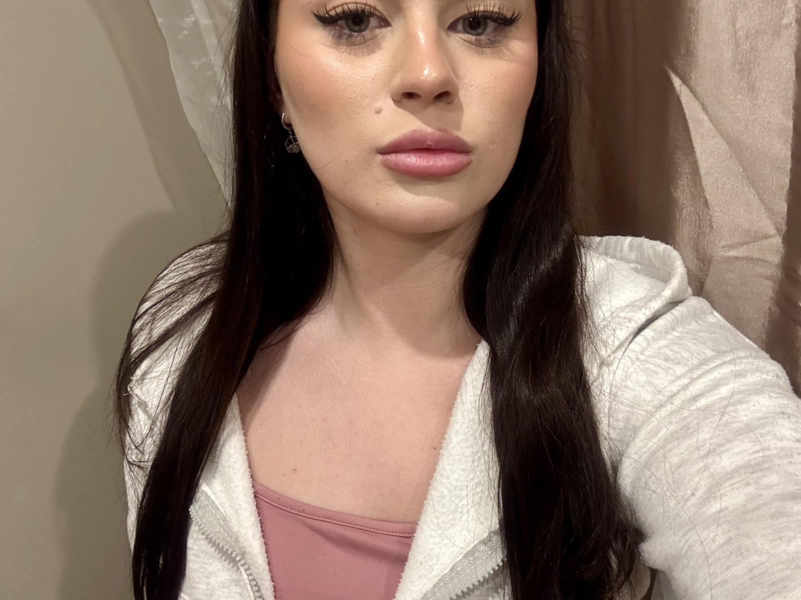 Mignon Cassel hat langes, dunkelbraunes Haar, helle Haut und pinken Lippenstift. Sie trägt eine weiße, mit einem Reißverschluss versehene Kapuzenjacke über einem pinken Oberteil, wodurch ihre kleinen Brüste sichtbar sind. Ihre Augen sind leicht nach oben geneigt..