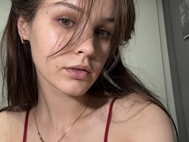 Nikoletes has light skin, brown eyes, and a small mole above her slightly parted lips. She's wearing a red strap, with long brown hair covering one eye.