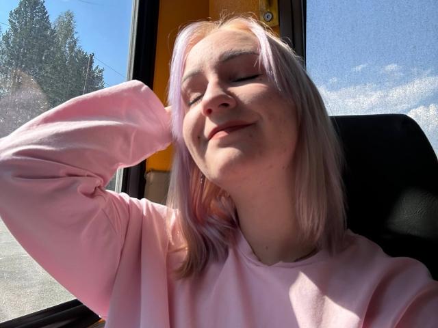 MacyGillyard with pink hair, eyes closed, smiling, wearing a pink sweatshirt, hand behind head, sunlight on face, sitting near a window, clear blue sky outside.