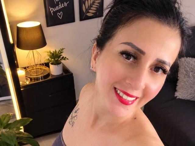 AgathaBrunner with dark hair in a messy bun, red lipstick, and a tattoo on her left shoulder, smiling at the camera. Her tits are slightly visible.