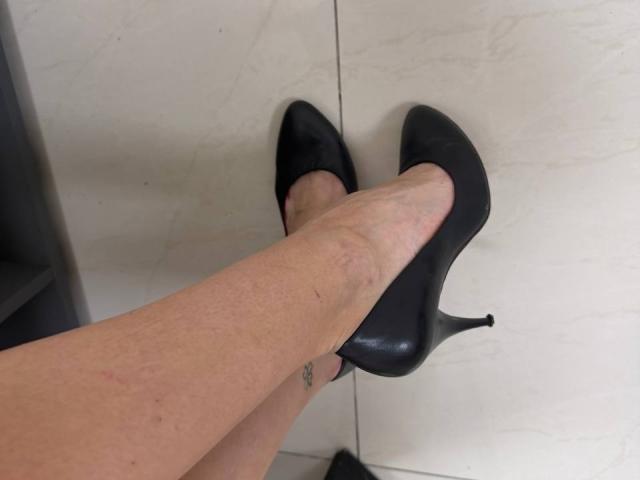 MoonLiza's pale legs in black stiletto heels, showing a small tattoo on her ankle, standing on white tiled floor. Her legs are slightly crossed at the ankles.