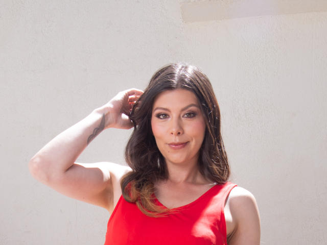 KarlaMazuera with wavy brown hair, wearing a red tank top, raising her right arm, showing a tattoo on her forearm, smiling at the camera.