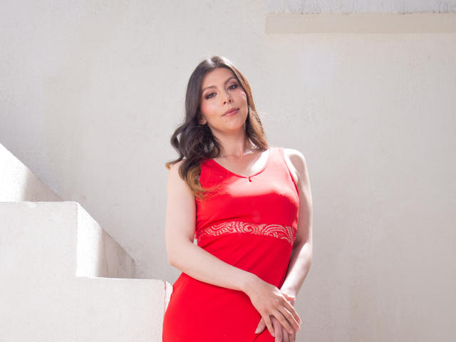 KarlaMazuera in a red dress, standing with hands on her belly, has long brown hair, light skin, and a gentle smile, showing her tits and ass in bright sunlight.